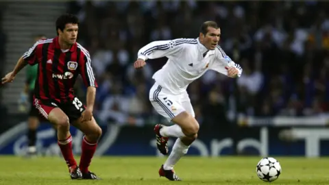 Getty Images Two footballers in game one wearing all white kit the other wearing black and red striped, the ball is ahead of them it is white with black stars on it