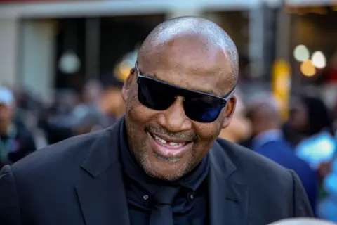 Dwayne Senior / Getty Images Gayton McKenzie mengenakan setelan jas, dasi, dan kemeja All Black, sambil mengenakan kacamata hitam saat ia menghadiri pidato negara bagian di Cape Town, Afrika Selatan. 