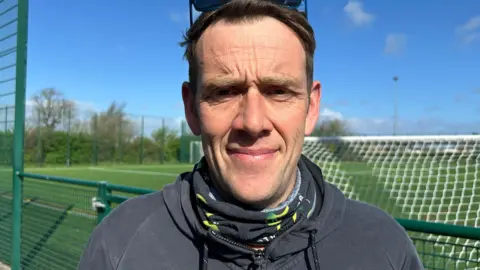 A man wearing a neck scarf and a zip up hoodie is standing in front of a green football training pitch with goals visible in the background. The sky is blue.