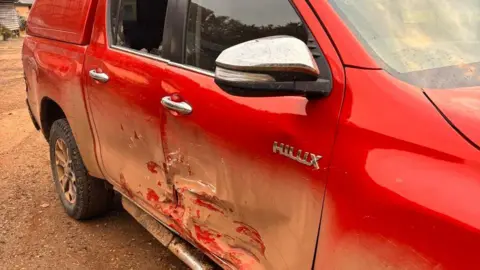 Berikan pickup Hilux merah, kendaraan yang terkenal kuat, dengan pintu depan dan belakang yang berkerut, sasis bengkok, dan jendela yang hancur.