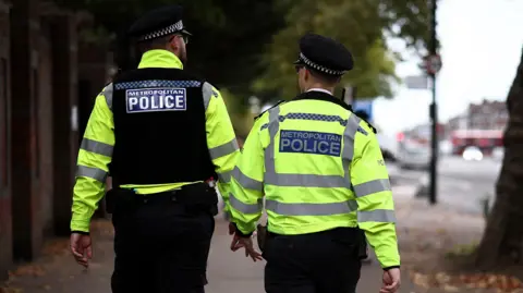 Two Metropolitan Police officers are walking forwards along a road with their backs to the camera. They are wearing high-visibility yellow jackets with the words "Metropolitan Police" on the back.