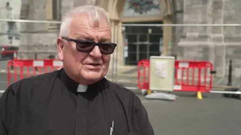 A middle-aged white an in clerical clothing and sunglasses stands outside a church entrance that is surrounded by temporary safety barriers and road closure equipment.