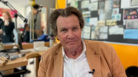 A man with brown hair, centre-parted and swept back, is looking into the camera. He has a white shirt on with a tan jacket over it and a small microphone clipped to one lapel.