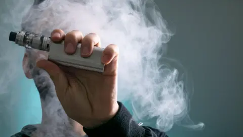 A silver vaping device being held close to the camera. A man's face is obscured by the device and a cloud of vapour.