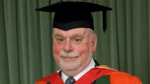 Professor Sir James Fraser Stoddart, pictured wearing a graduation cap and red robe. He has a grey goatee.