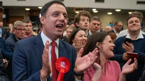 Anas Sarwar claps his hands as he celebrates Scottish Labour's by-election victory with a group of party activists.