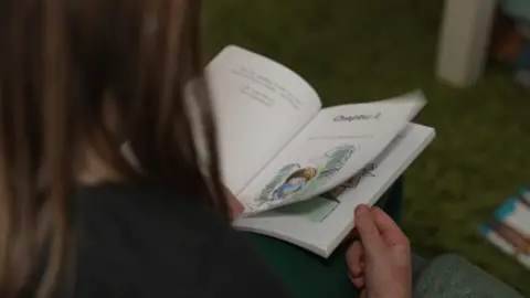 The picture shows a child sitting on a soft, green carpeted floor and reading a small illustrated book. The child is holding the book open on their lap, turning a page to a chapter that includes a colourful drawing of a character. Only the back of the child’s head, shoulders and hands are visible, so you cannot identify them. 