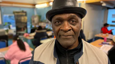 Lloyd Russell at the family history class, looking into camera. He is wearing a black pork pie-style hat and a cream-coloured jacket