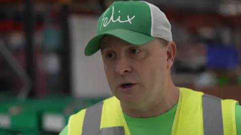 A man in a high-vis tabard wearing a cap with the word Felix on the front talks to a reporter in a distribution warehouse. 