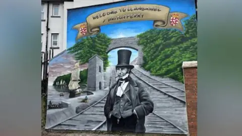 LDRS A mural of Isambard Kingdom Brunel on the side of a building. He wears a top hat, waistcoat and coat, with a cigar in his mouth, and stands in front of a railway track and a bridge. Above the bridge are the words 'welcome to Llansawel/Briton Ferry'. 