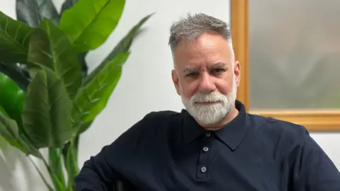 A man with a navy blue long sleeved polo shirt and grey hair. To the left of the photo there is a large green plant and to the right there is a framed picture.