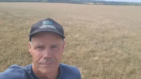 Duncan Palmer, a middle-aged man wearing a baseball cap is stood in a field of yellow grass.