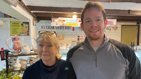 A man and a woman standing in a cafe. The man is wearing a grey and black half zip sweater, the woman is wearing a black roll neck top with short blonde hair and black glasses sitting on top of her head