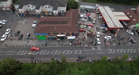 Virtual Towns An aerial view of crowds gathering on the Stewartstown Road. People can be seen gathering in a car park beside shops. There are houses at the rear of the picture and a road in the foreground.
