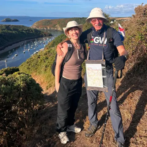 Andy Hockey John Parry and Kyla Norrie stand smiling at the camera. They are both wearing white sun hats, and have their arm around each other's shoulders. John is wearing a 'Grandad's Great Walk' charity t-shirt, while Kyla wears a brown vest. The pair are standing on a footpath in north Pembrokeshire, with a view of hills behind them.