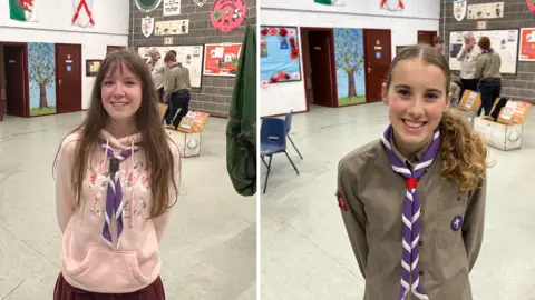 BBC/RICHARD EDWARDS These pictures show two explorer scouts, both called Emily, at a weekly unit meeting in Copmanthorpe, York. Emily on the left has long brown hair and is waering a pink hoodie and a purple and white neckerchief. Emily on the right has her brown hair in a ponytail and is wearing a khaki-coloured scout uniform shirt and the same coloured neckerchief. They are both smiling and are standing in the hall used by the local scouts for their regular get-togethers.