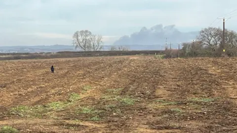 Smoke can be seen billowing in the sky in the distance. The photo is taken in a large brown/muddy field.