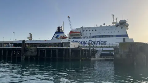 BBC An image of a blue and white Brittany Ferries boat at the Guernsey Port, which has stairs and pillars leading up from the ocean and 'Welcome to Guernsey' written on the side. 