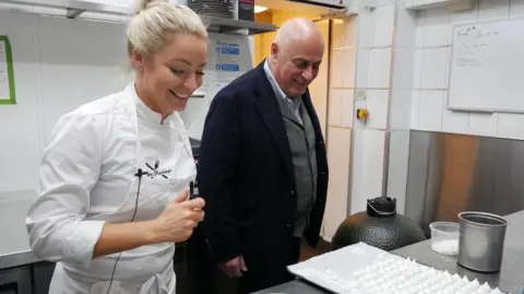 Claire Hutchings Claire Hutchings, wearing a white chef top, and with blonde hair pulled back in a bun, looks at a metal worktop, standing next to a shaven-headed man in a kitchen.