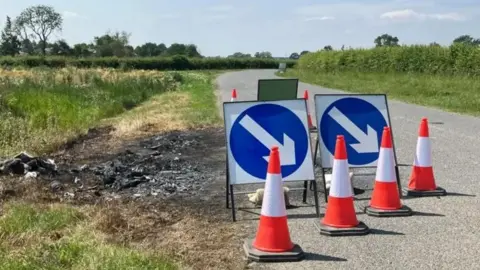 A road in the countryside with burnt out grass by the side where the car was found and traffic cones and signs with arrows on placed next to it