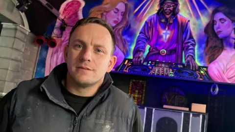 BEN SCHOFIELD/BBC Nathan Murdoch looking directly down and the camera and standing in front of a stylised spray-painted mural. Nathan is wearing a blue gilet-style coat, which has marks of paint on it, and a black long-sleeved top. He has short, light brown hair. The mural behind him shows a bearded man in robes operating a DJ's set of turntables, with female characters on either side of him. Nathan is lit from the right, with one side of his face brighter than the other. 