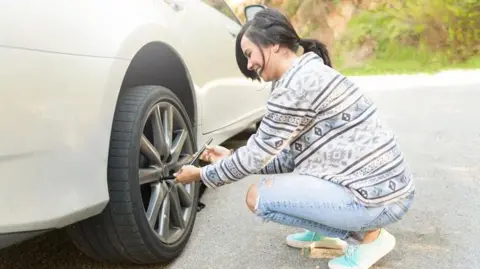 A young woman stranded on the side of the road deals with a flat tyre. She is crouching by a wheel and holding a gadget. She is wearing jeans and a jumper and trainers, with her hair in a ponytail.