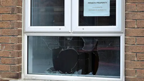 A smashed window is in-between two sides of a brick wall. The window is white. It has a sign on the top right segment saying private property. 