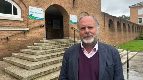 Julian Brazil, Liberal Democrat leader of Devon County Council in a pink shirt, burgundy sweater and blue blazer