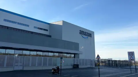Vue extérieure de l'entrepôt Amazon à North Ferriby. C'est un grand bâtiment blanc avec le logo Amazon sur un côté et un grand parking devant. Le ciel est bleu. 