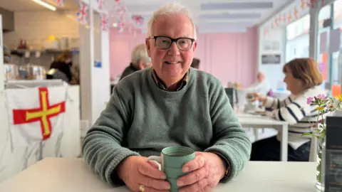 David Savident pictured wearing glasses and a green sweater sits at a table in a bright café, smiling while holding a light green mug. The café has white walls, pink accents, and several Union Jack flags hanging from the ceiling. Other patrons sit at nearby tables, and a counter with kitchen equipment and a white flag featuring a red cross with a yellow border is visible in the background.