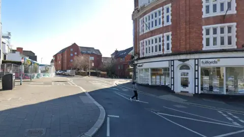 A man walks across the entrance to Gibbon Lane with a shop front on the right side and flats further up the street. 