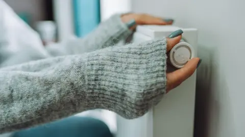 Getty Images A woman places her hands, which are semi-covered by her grey cardigan, turns a dial on a radiator. The image is a close-up of the womans hands.