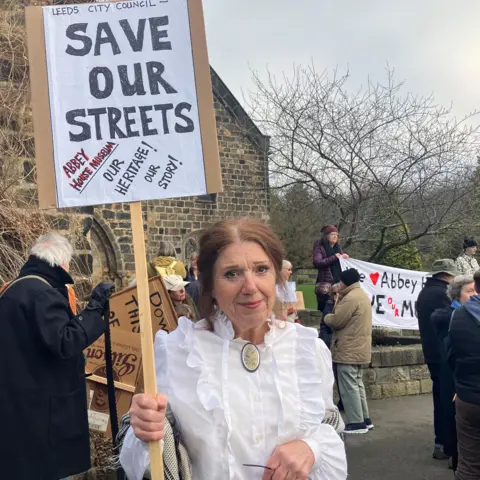 BBC/Corrine Wheatley A woman wearing Victorian costume - a white blouse with frills and a neck clasp - holds a placard reading "Save our Streets". She is standing with fellow protesters in front of a museum building.