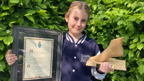 A young girl smiles into the camera holding a plaque in her left hand and a foot-shaped trophy in her right hand. 