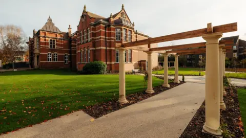 A large Victorian red-brick building stands on lawns, there is also a pathway with a pergola across. 