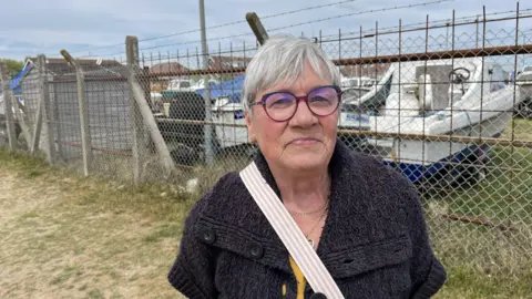 Phil Harrison/BBC Marlene Hubbard stood beside a fence with some small boats in the background.