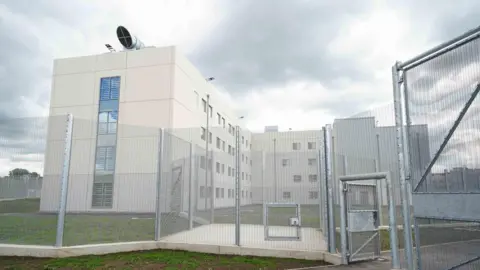 HMP Fosse Way, a white prison with silver fencing and gating surrounding it