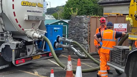 BBC The image shows two large tanker lorries connected by hoses in a street, with a worker in high-visibility clothing standing between them near traffic cones, likely transferring liquid at a wastewater or drainage site.