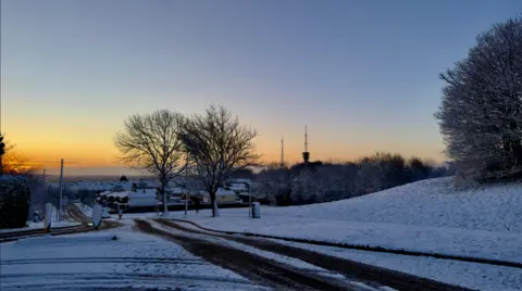 A road which is partially covered in a light layer of snow, which is worn off in the centre by tyre tracks. Snow can be seen on an embankment in the right of the image, the photo has been taken at dawn and the sun can be seen rising in the distance