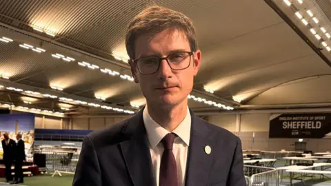 A man stands in a leisure centre where tables are in the background for the vote count. He has a navy suit, burgundy tie, brown short hair and black square glasses. He has a neutral expression but looks sad.