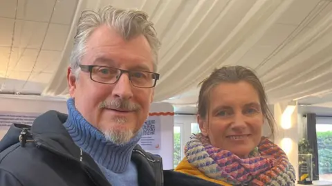 Connor Bennett/BBC A couple smile at the camera in a head and shoulders image of them both, standing side by side. The man, to the left, has blue eyes, grey hair and black framed spectacles, and is wearing a blue polo neck jumper and navy jacket. To the right, the woman has blue eyes, brown hair tied back, a multi-coloured scarf and dark blue jacket.