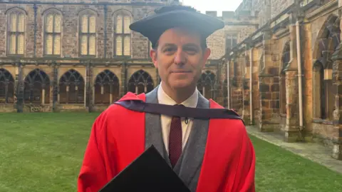 Matt Baker wearing a red graduation cloak, on top of a grey blazer with a maroon tie, holding up a two sided folder which holds his certificate and documents. 