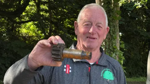 Luke Deal/BBC A head and shoulders image of David wearing a grey top with the Battisford & District club logo on it. He is holding up a large metal cog with a metal rod attached to it. He is standing outside in front of some trees.