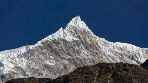 A panomanic view of the Langtang Lirung peak. The snowy summit is seen against a clear blue sky.