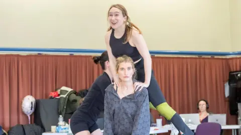 Johan Persson A woman with long blonde pigtails jumps on to the back of another woman with tied up blonde hair. They are in a theatre rehearsal room
