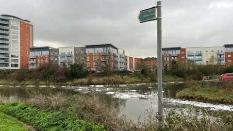 Connor Bennett/BBC A river with spots of white foam on it during a grey and cloudy day. Blocks of flats can be seen in the distance. A sign detailing the footpath next to the river stands in front of the river.
