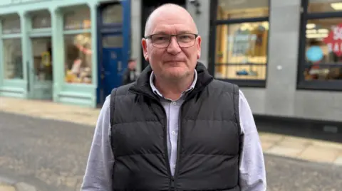 John Wellwood looking straight at the camera. He is wearing a black gilet over a white shirt.