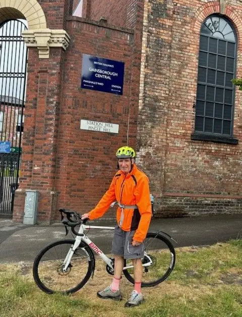 John Whitbread A man wearing an orange top and grey shorts standing with a white bicycle close to the arched entrance to a retail park.