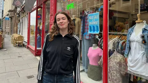 A woman wearing a black zip up Adidas jacket. She has dark brown curly hair. In the background is her shop which has a red front and 