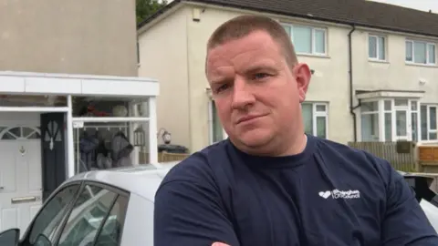 BBC A man with short brown hair and a blue tshirt with his arms folded in front of a white car and some white-painted houses